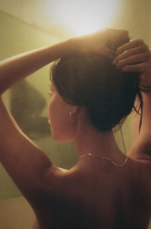 Adult female in warm-toned indoor setting, touches her hair. Gold necklace and hoop earrings are visible in a candid shot.
