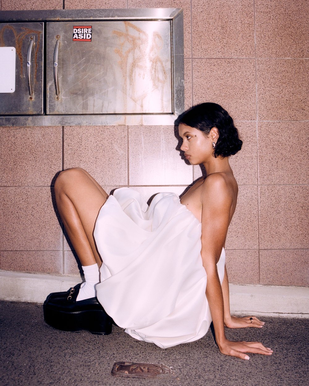 Young woman in a white dress poses in a tunnel, wearing platform shoes and white socks, lit by natural light, with a tiled wall.