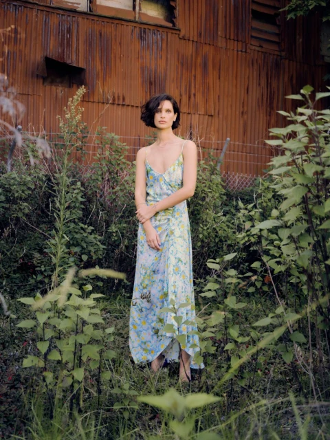 Woman in floral dress stands in lush outdoor setting near weathered building, capturing a blend of fashion and natural elements.