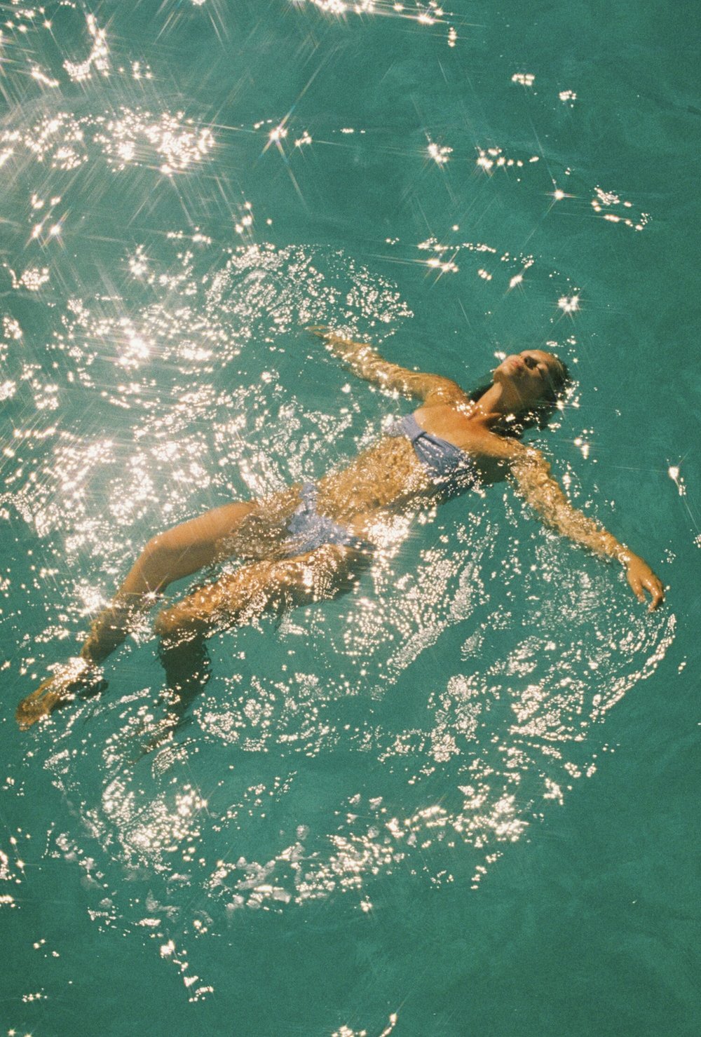Woman in a blue bikini floats on her back in bright, shimmering, turquoise water with her arms extended at her sides on a sunny day.