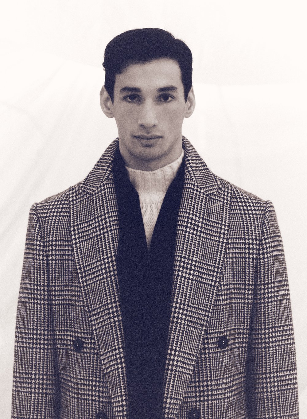 Portrait of a stylish adult male wearing a patterned overcoat with a neutral-colored turtleneck and dark scarf against a plain background.
