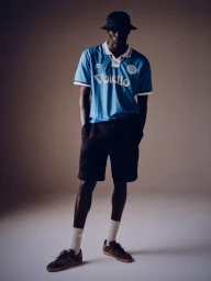 Black man standing wearing a blue football jersey, dark shorts, bucket hat, and sneakers in a studio