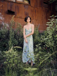 Woman with short dark hair wearing a floral slip dress standing outdoors in overgrown weeds before a rusty metal wall