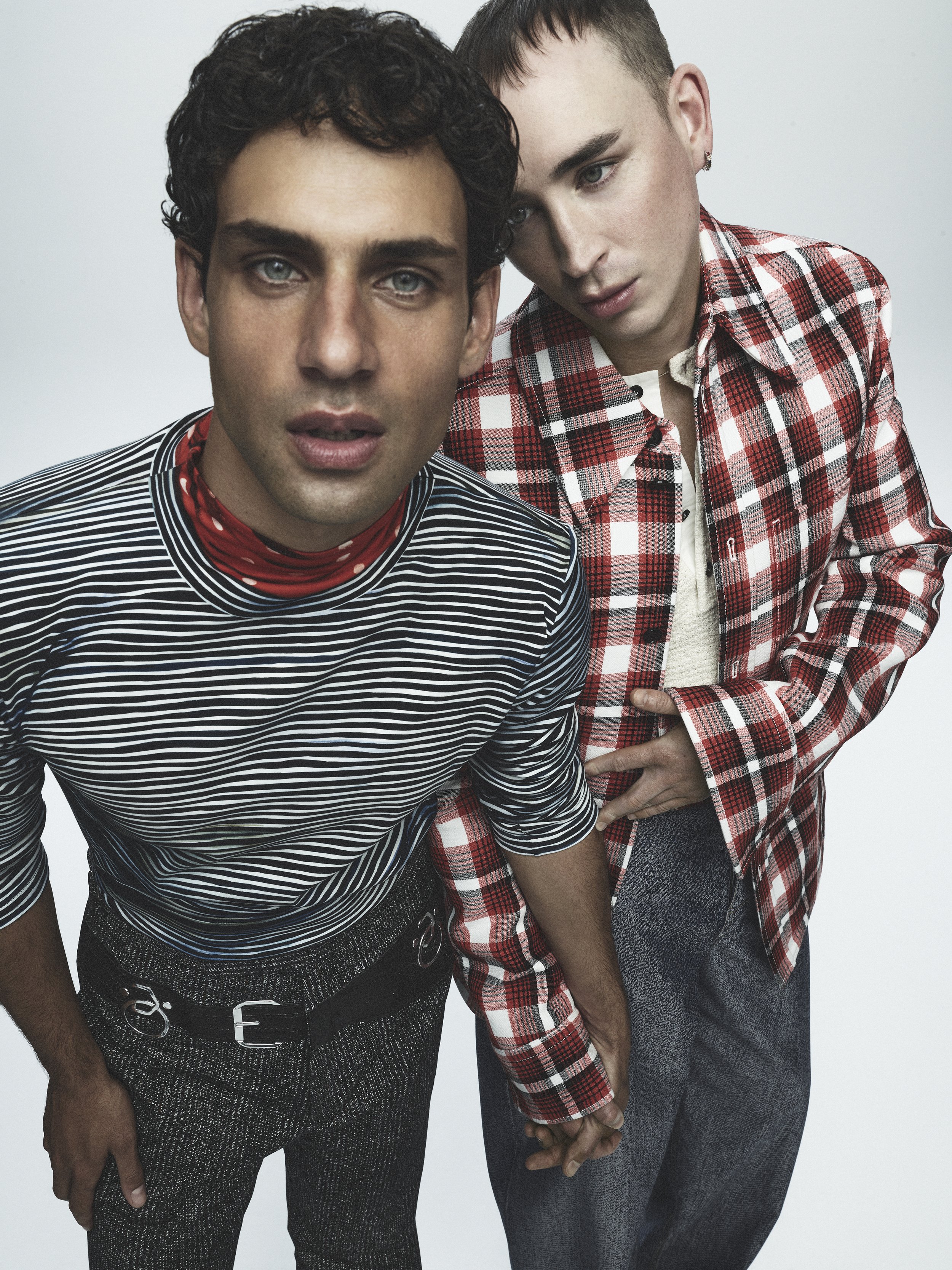 Two men in casual attire pose indoors, one in a striped shirt, the other in a red plaid shirt, holding hands in a modern portrait.