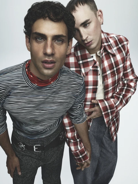 Two men in casual attire pose indoors, one in a striped shirt, the other in a red plaid shirt, holding hands in a modern portrait.