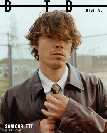 Sam Corlett adjusting his tie, wearing a brown leather jacket and collared shirt styled by Thomas Townsend.