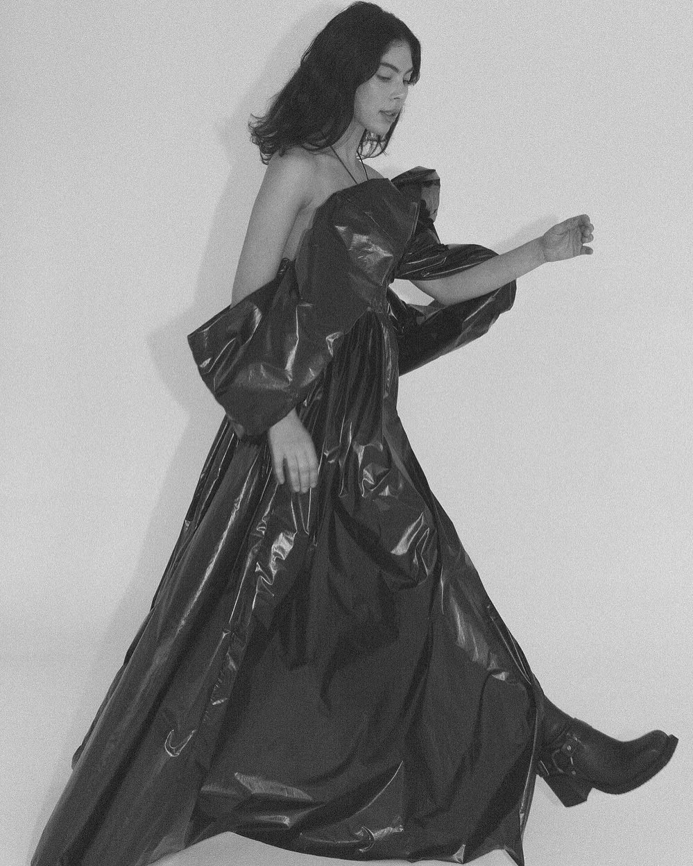 A person with dark hair walks sideways wearing a shiny black gown and matching boots against a white background in a studio-style setup.