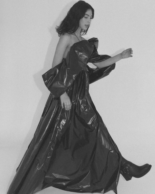 A person with dark hair walks sideways wearing a shiny black gown and matching boots against a white background in a studio-style setup.