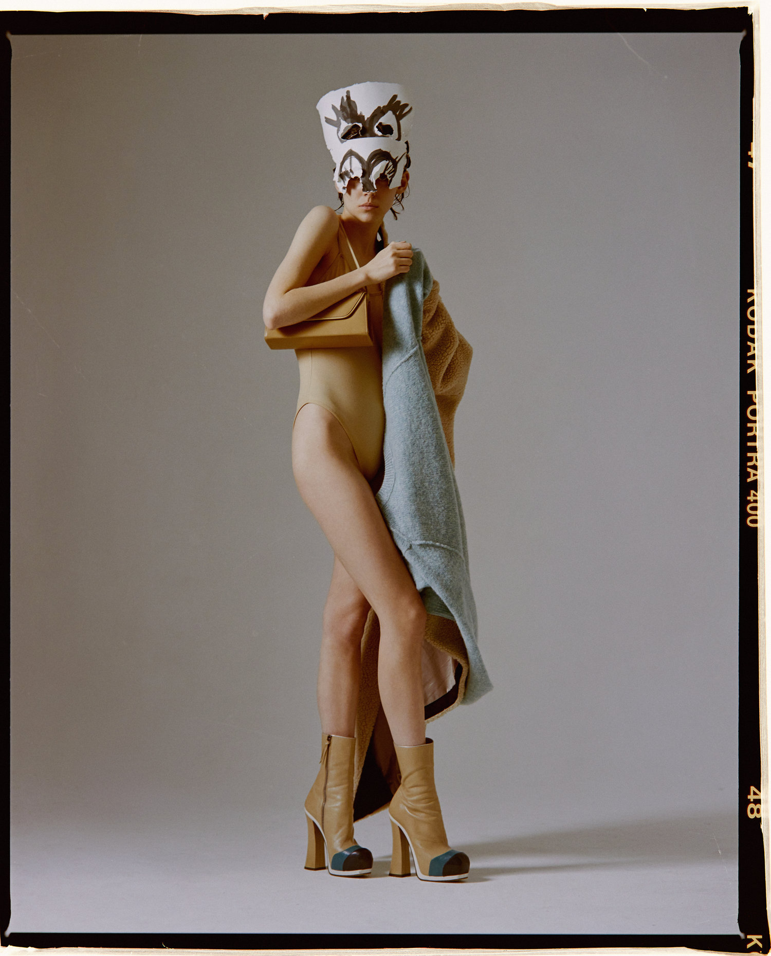 A fashion shot featuring an adult female model in a bodysuit with a decorative hat, boots, and a purse in a photo studio setting.