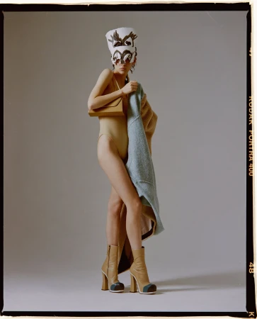 A fashion shot featuring an adult female model in a bodysuit with a decorative hat, boots, and a purse in a photo studio setting.