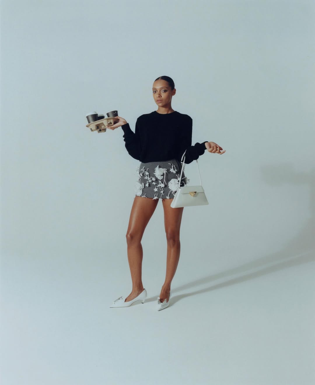Elegant woman in a black top and floral skirt balances a tray of coffee and a purse in a light studio setting. 