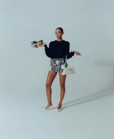 Elegant woman in a black top and floral skirt balances a tray of coffee and a purse in a light studio setting. 