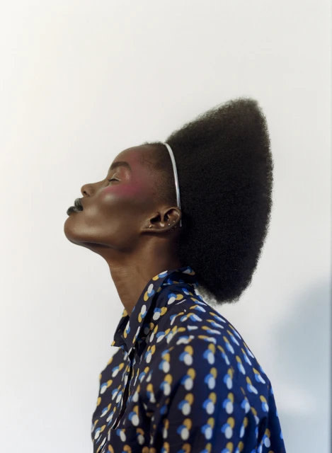 Profile shot of an African-American woman with a stylish afro and a patterned button-down shirt against a stark white background.