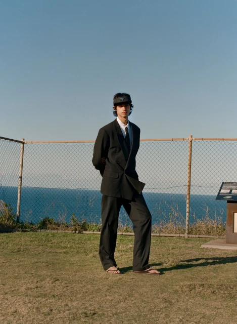 An adult male stands on a grassy hill overlooking the ocean, wearing a black suit and visor, blending formal wear with a natural setting.