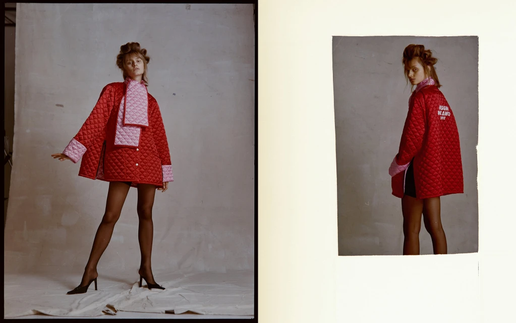 Fashionable adult woman in a red, quilted jacket, pink scarf, and black hosiery, posing in a studio setting with a grey background.