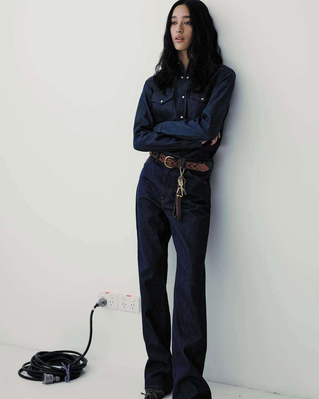 A woman with long dark hair poses in a dark denim shirt and jeans against a white wall. Cord and wall outlets are visible.