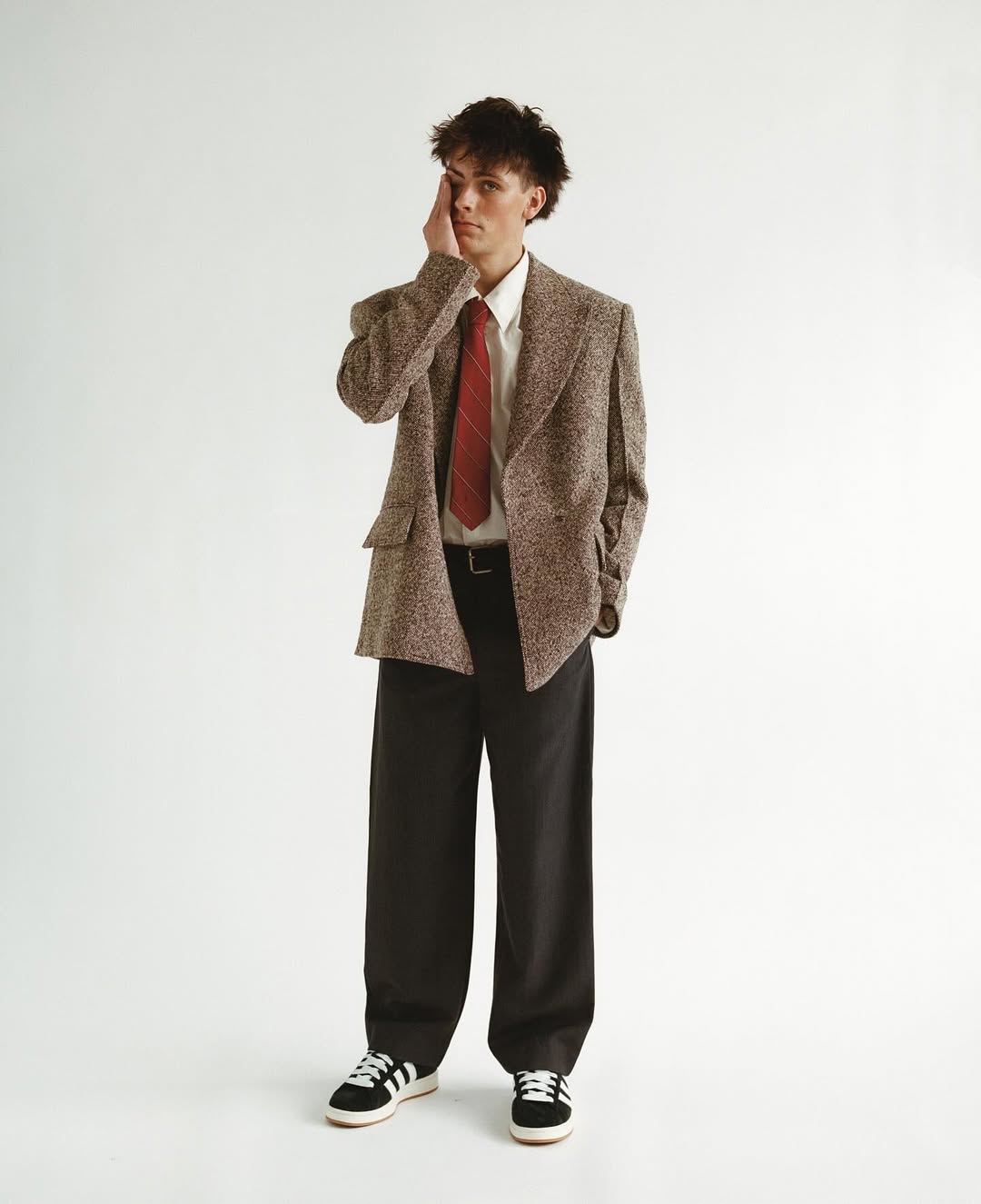 Full-length studio shot of a young adult man in a brown tweed blazer, white shirt, red tie, and black pants posing with hand on face.