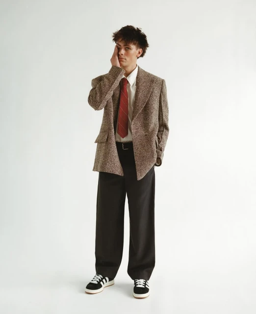 Full-length studio shot of a young adult man in a brown tweed blazer, white shirt, red tie, and black pants posing with hand on face.