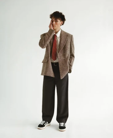Full-length studio shot of a young adult man in a brown tweed blazer, white shirt, red tie, and black pants posing with hand on face.