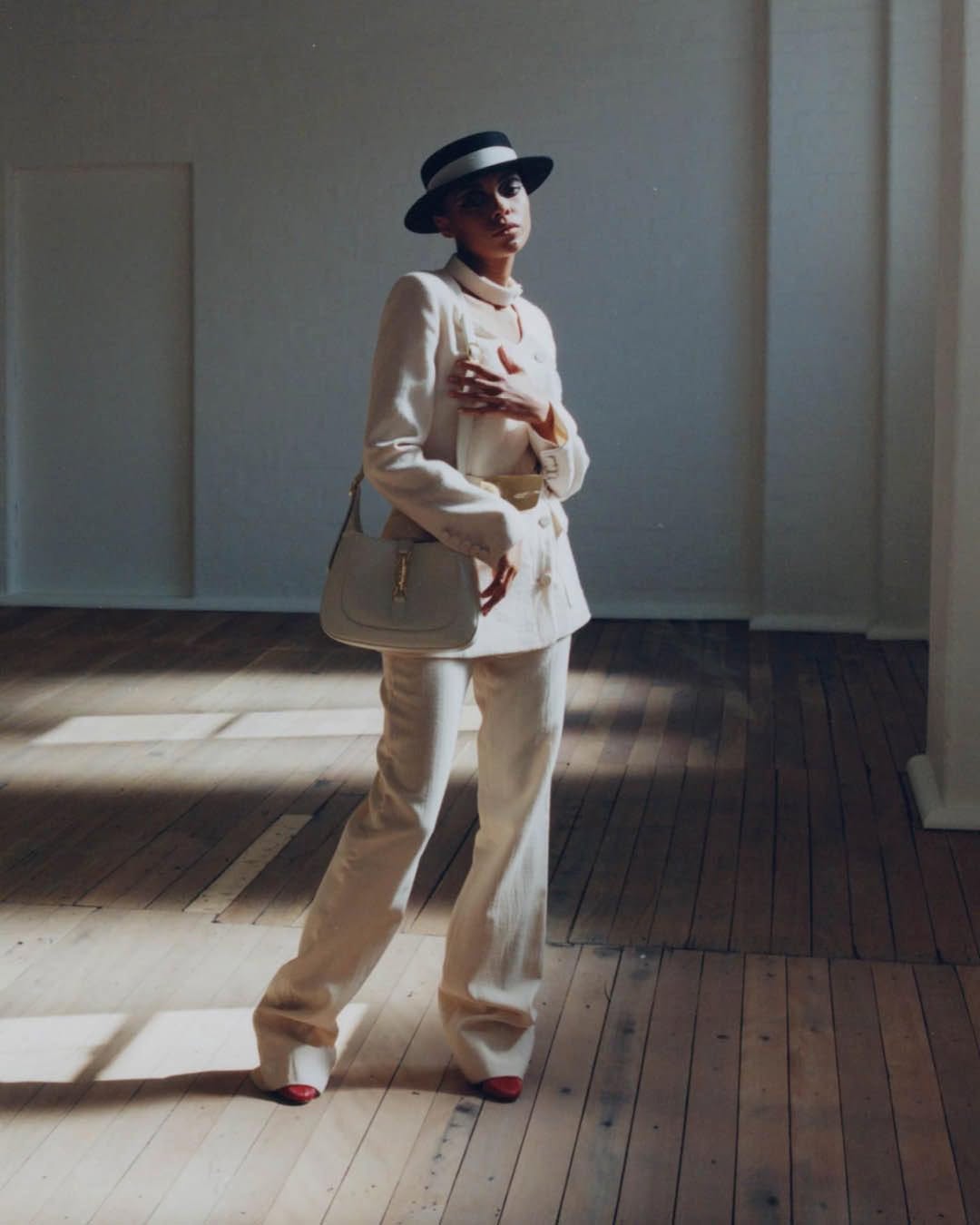 Elegant woman in a cream suit and hat poses in a room with wood floors and sunlight, carrying a matching cream handbag.