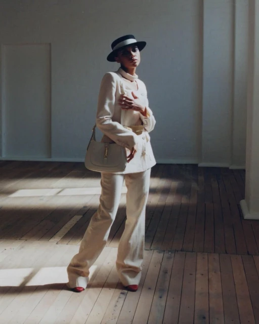 Elegant woman in a cream suit and hat poses in a room with wood floors and sunlight, carrying a matching cream handbag.