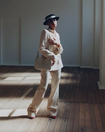 Elegant woman in a cream suit and hat poses in a room with wood floors and sunlight, carrying a matching cream handbag.