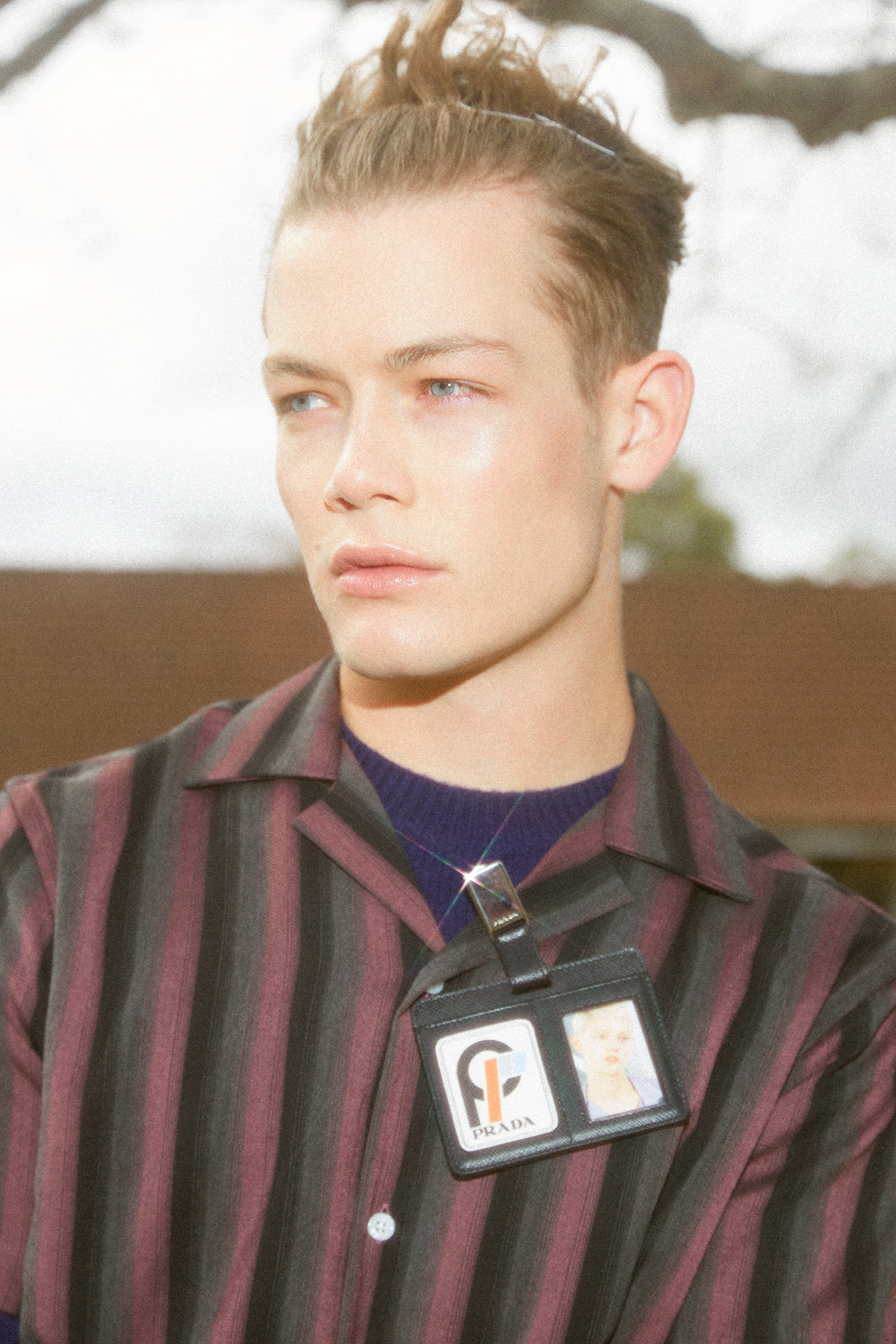 Close-up portrait of a stylish adult male model wearing a striped shirt and Prada identification badge outdoors on an overcast day.