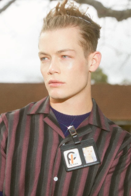 Close-up portrait of a stylish adult male model wearing a striped shirt and Prada identification badge outdoors on an overcast day.