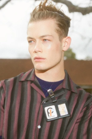 Close-up portrait of a stylish adult male model wearing a striped shirt and Prada identification badge outdoors on an overcast day.