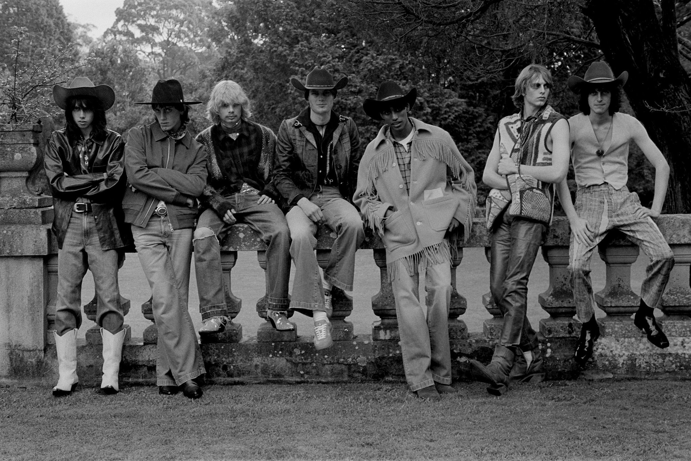 Black and white shot of seven people wearing hats and Western-style clothing, sitting outside on a stone ledge with trees in the background.