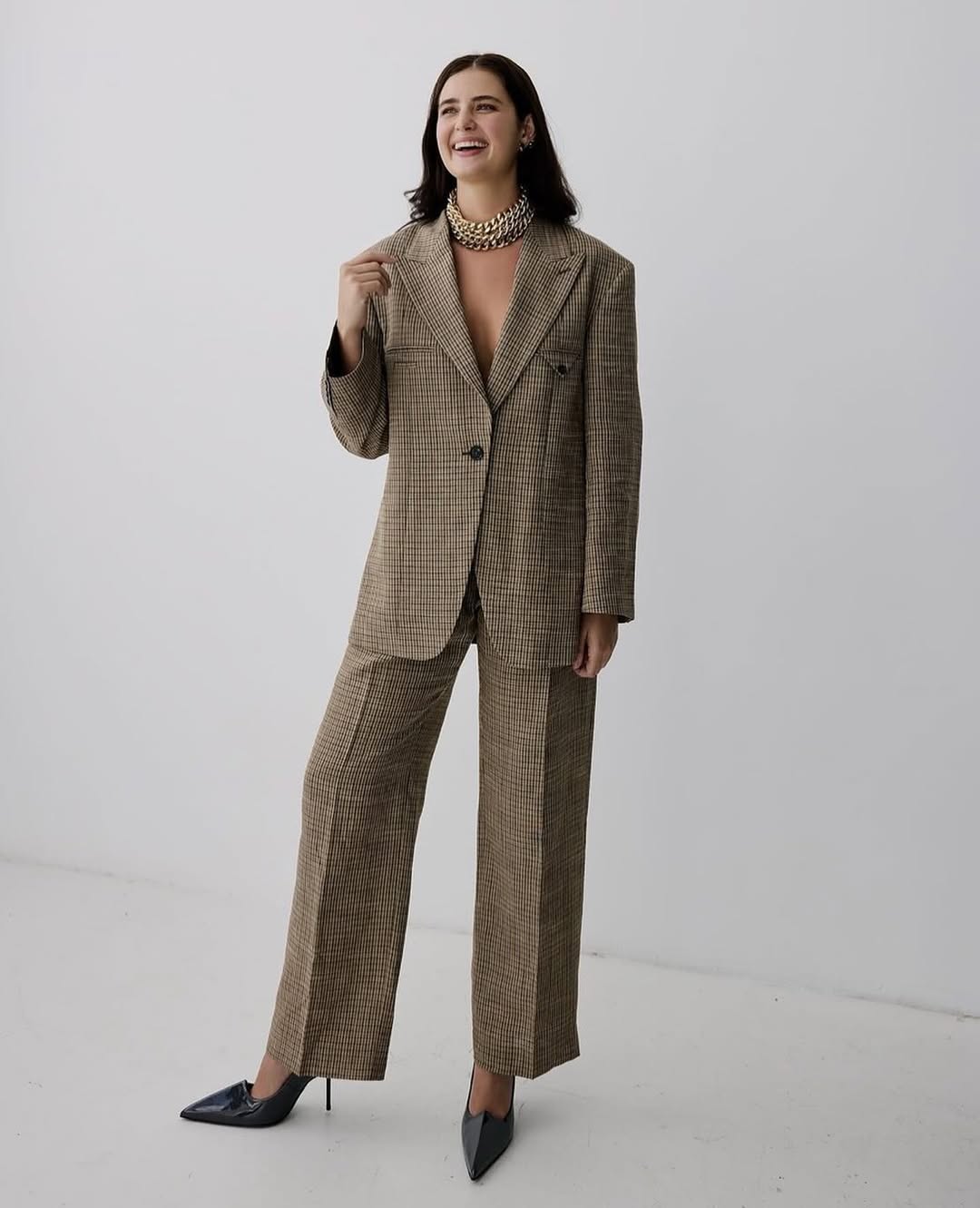 Smiling brunette woman in a brown plaid suit and black heels stands in front of a white wall looking off-camera.