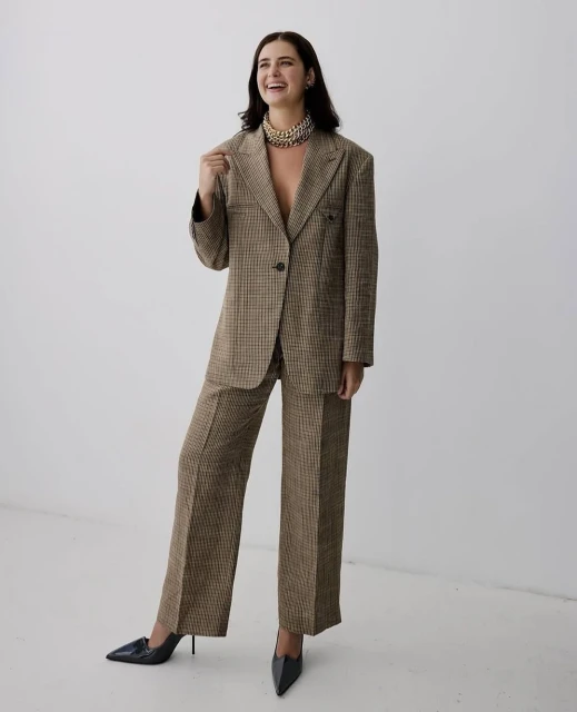 Smiling brunette woman in a brown plaid suit and black heels stands in front of a white wall looking off-camera.