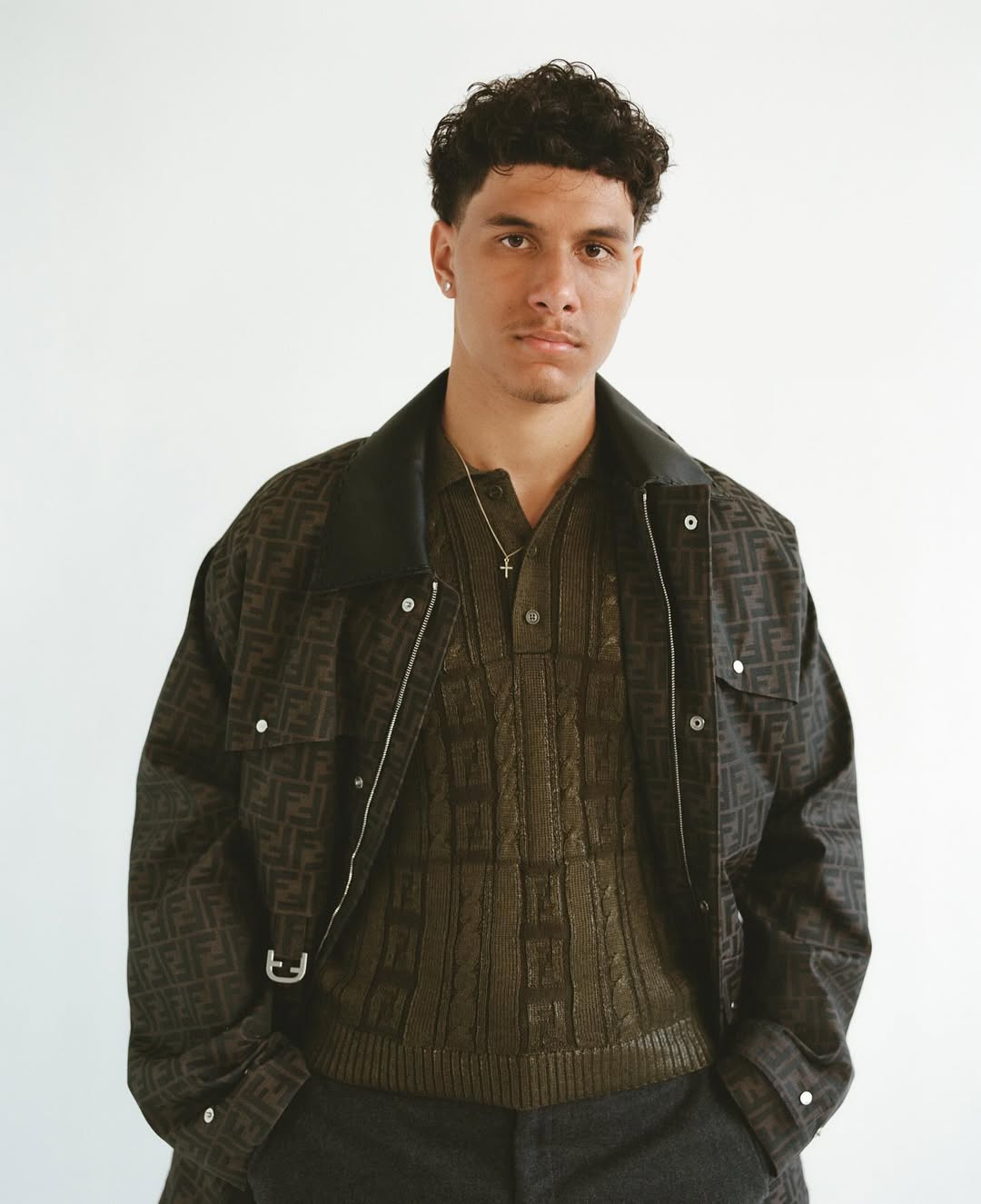 Man in patterned jacket and shirt poses in a studio. Portrait of a young adult male wearing olive green knitwear and a patterned jacket.