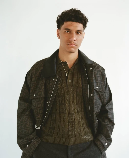 Man in patterned jacket and shirt poses in a studio. Portrait of a young adult male wearing olive green knitwear and a patterned jacket.