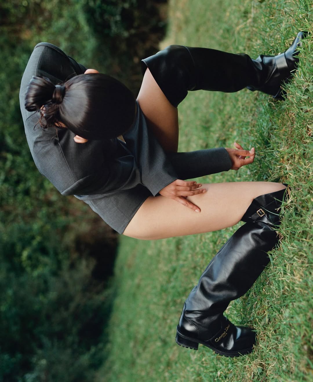 Overhead shot of a woman in a grey jacket and thigh-high boots sitting on green grass in an outdoor environment with soft natural light.
