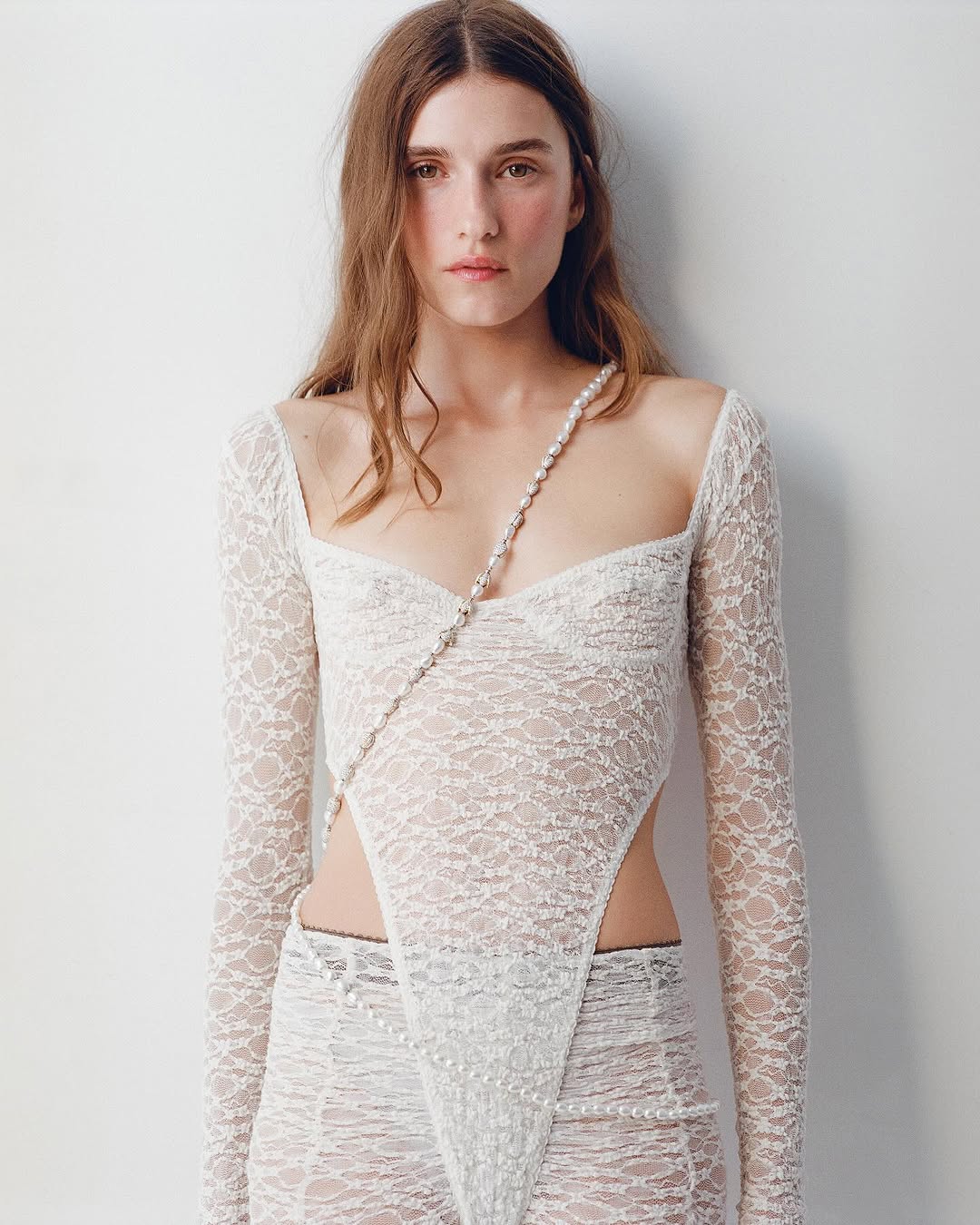 A fair-skinned fashion model with long brown hair poses in a white lace top and pants, accentuated by a pearl necklace, against a white backdrop.