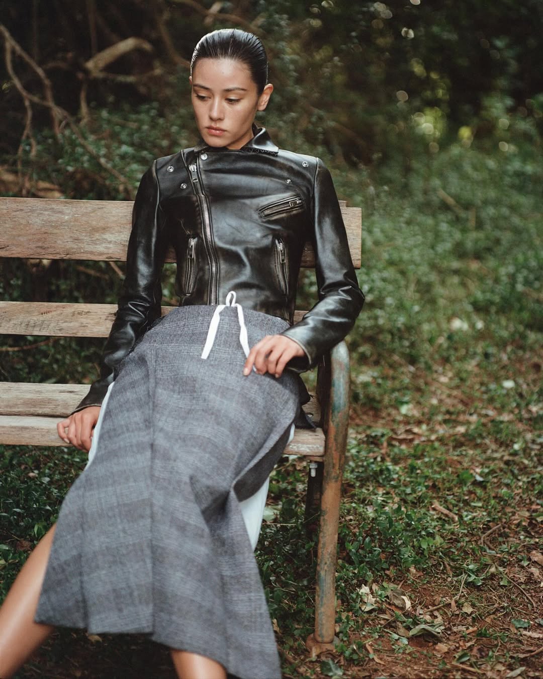 Woman in black leather jacket and grey skirt sits on a park bench surrounded by greenery, posing and looking down in a medium shot.