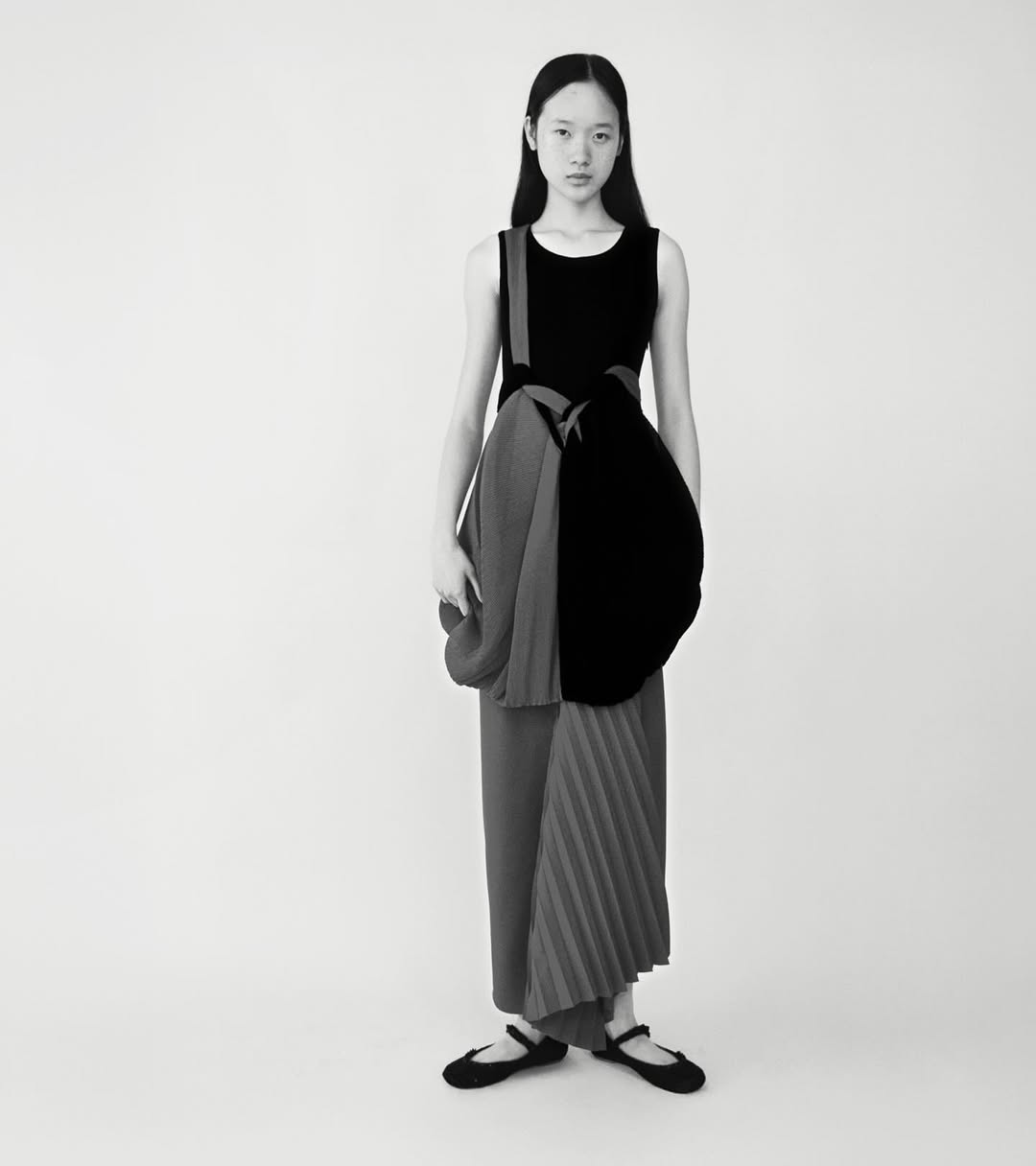 Monochrome fashion shot of a slender Asian woman in an asymmetrical black dress with pleated detail against a plain background.