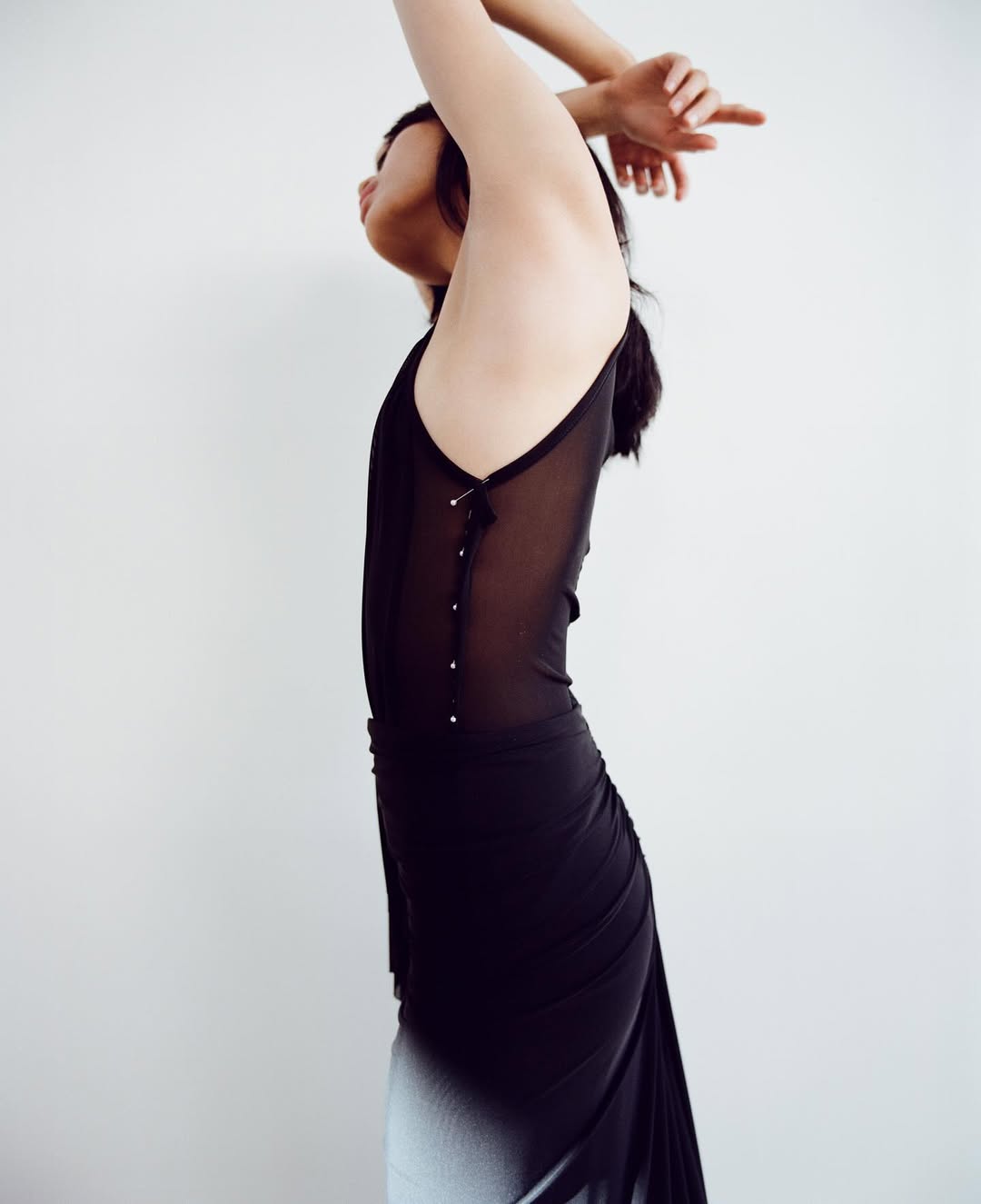 Slender woman in semi-sheer black dress with a gradient hem, arms raised against a plain white background in a fashion-forward shot.