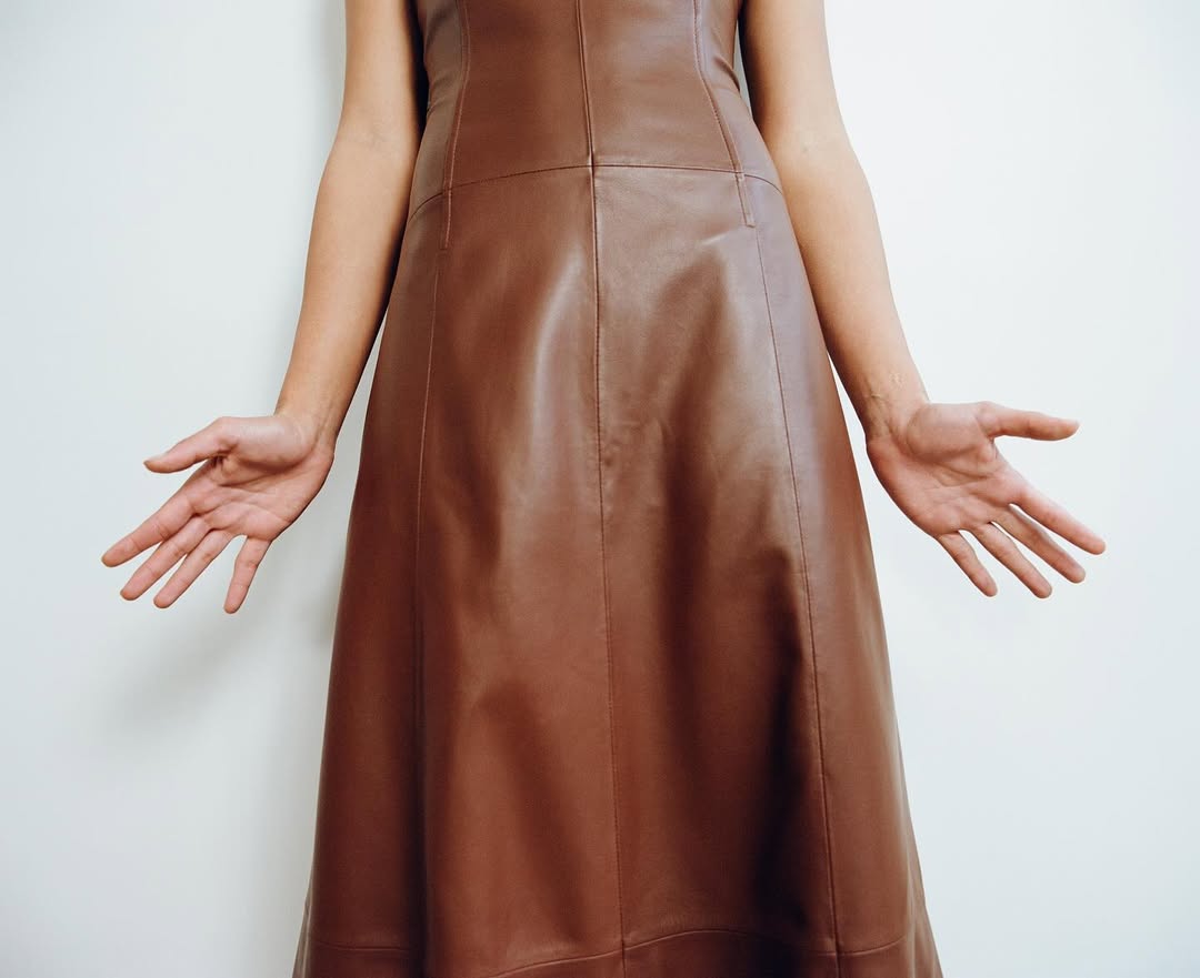 Close up of a woman's torso wearing a brown leather dress with her arms outstretched against a plain white background, fashion-forward.