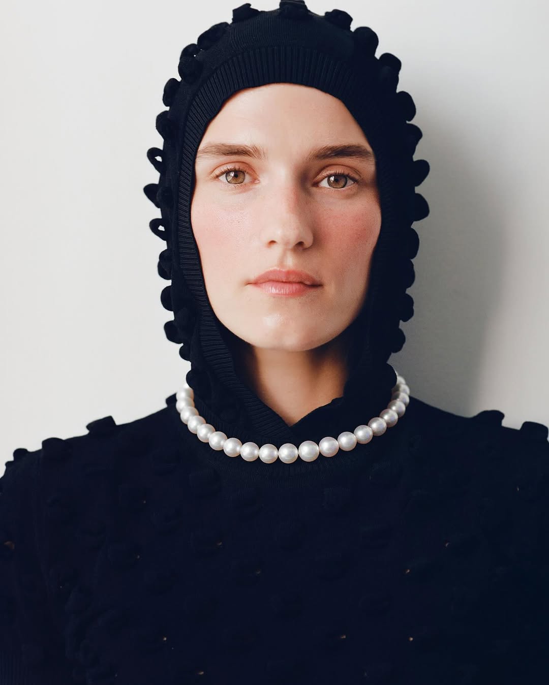 Portrait of a woman wearing a black knitted balaclava with frills and a pearl necklace, set against a plain white background.
