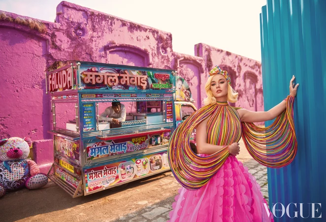 Katy Perry poses in front of a dessert cart and pink wall with an Indian man inside the cart and blue columns beside her.