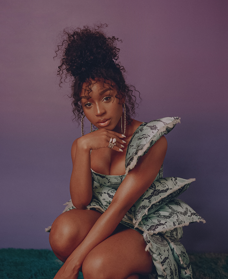 Portrait of a young Black woman with curly hair posing in a patterned dress against a gradient purple backdrop in a studio setting.