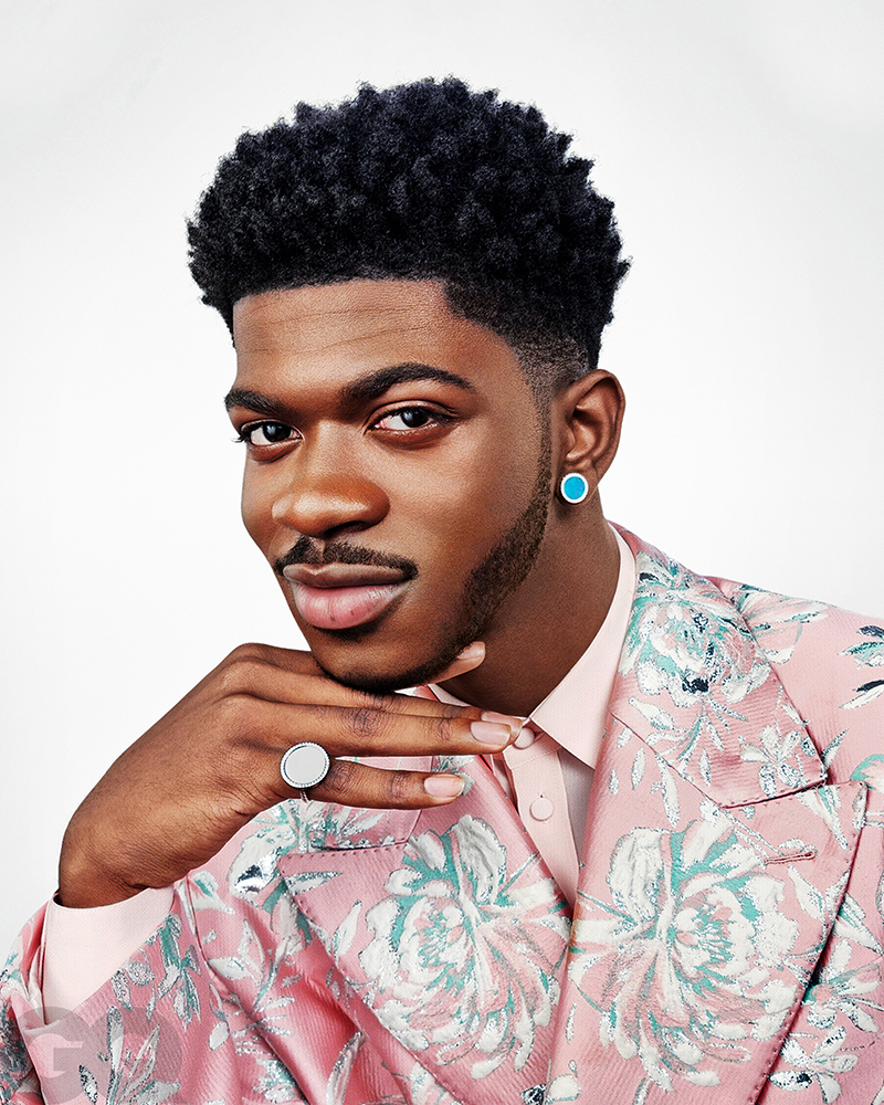 Close-up portrait of a Black man wearing a patterned pink blazer and turquoise earrings, with a faint smile, against a white backdrop.