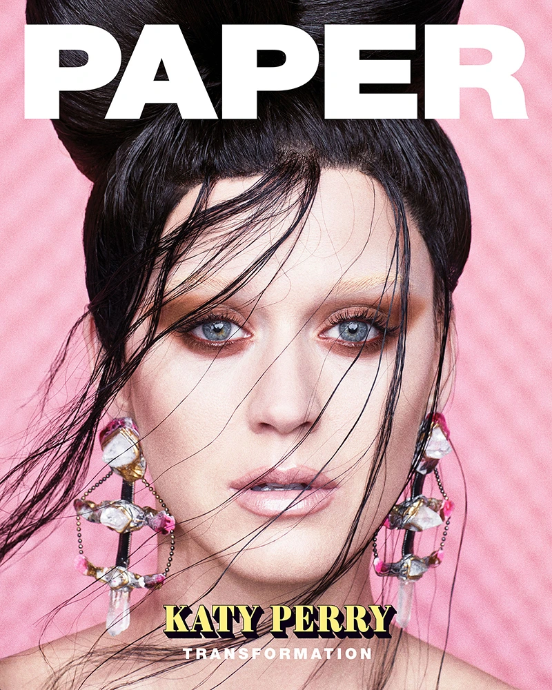 Close-up portrait of Katy Perry with dark wet-look hair and ornate crystal earrings against a pink textured background.