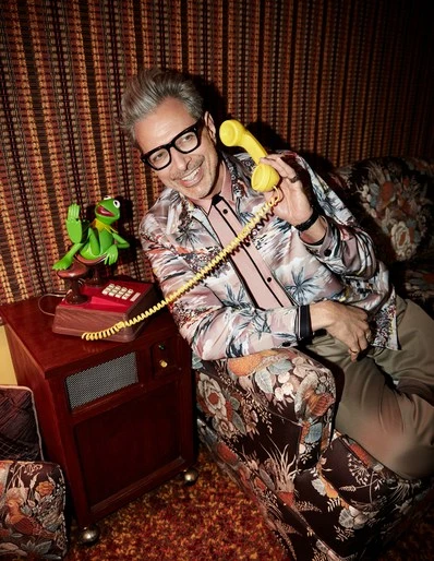 Jeff Goldblum sits on a floral sofa, holding a yellow telephone receiver next to a red phone with a Kermit the Frog figure.