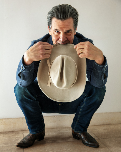 Adult male in denim jacket and jeans squats with cowboy hat, eyes on the camera, against a white wall, creating a classic portrait.