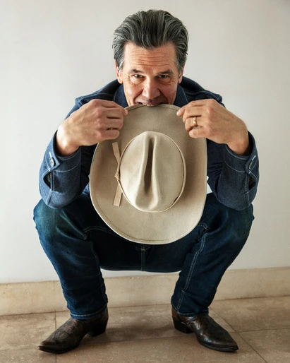 Adult male in denim jacket and jeans squats with cowboy hat, eyes on the camera, against a white wall, creating a classic portrait.