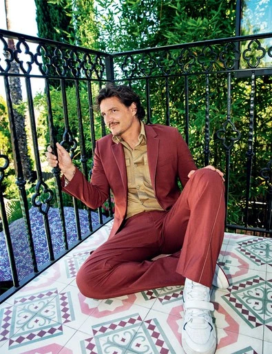 Pedro Pascal poses in a red suit on a patterned patio. He wears white sneakers and sits on a colorful tile surface with green plants.