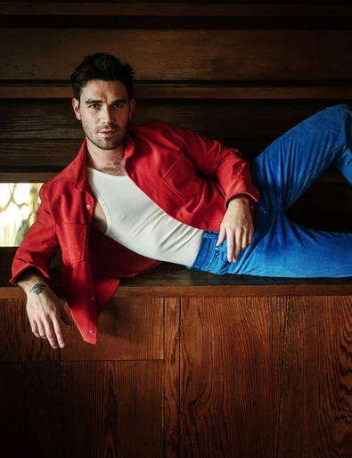 Man lounges indoors, wearing a red shirt and blue jeans, looking directly at the camera with a serious expression in soft natural light.